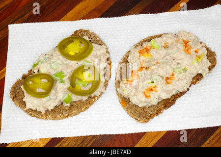 Aprire di fronte insalata di tonno pasto a sandwich realizzata con il sedano, la maionese, condimenti, jalapeno fette e la salsa calda sul grano intero pumpernickel pane di segale servita su un asciugamano di carta su un vassoio di legno Foto Stock