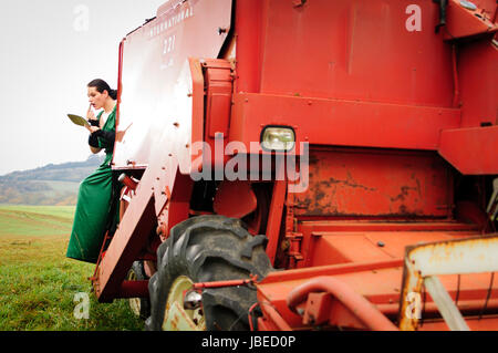 Erwachsene Frau im eleganten Abendkleid sitzt im Feld auf einem großen Mähdreschre und schminkt sich. Foto Stock