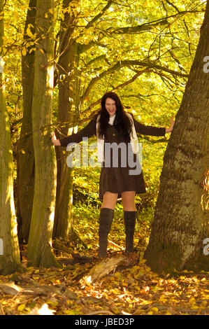 Junge übergewichtige Frau mit langen Haaren dunklen in erdfarbener Kleidung und braunen Stiefeln herbstlichen im Wald unterwegs. Foto Stock
