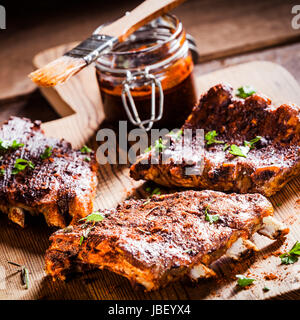 Tre porzioni di costolette alla brace in una marinata piccante condita con pepe e erbe aromatiche fresche di essere preparati in una cucina di paese con un barattolo di salsa di accompagnamento Foto Stock