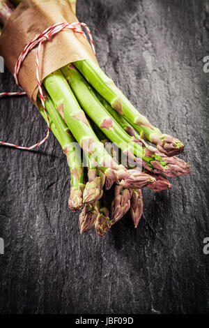 Fattoria fresco di punte di asparagi in un involucro marrone legati con spago giacente su una superficie strutturata di ardesia grigia con superficie di vignettatura e copyspace per il testo Foto Stock