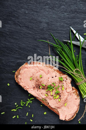 La preparazione di un delizioso sandwich teewurst spalmato su una fetta di pane di segale e guarnita con erba cipollina fresca tritata su una vecchia cucina ardesia contatore con copyspace Foto Stock