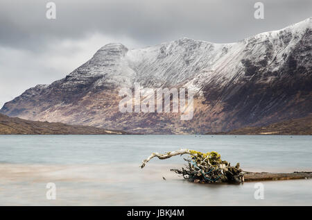 Loch Damh in Torridon, Scozia, con montagne innevate in lontananza e un vecchio albero carrello galleggiante nel lago in primo piano Foto Stock