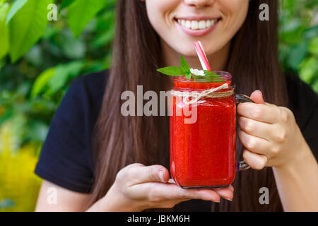 Primo piano della ragazza sorridente vetro azienda mason jar del frullato fragole nelle sue mani. La dieta, cibo vegetariano, uno stile di vita sano concetto. Donna di bere Foto Stock