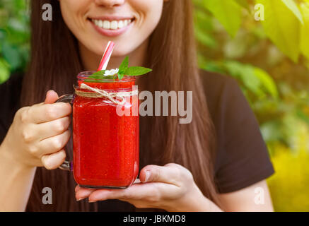 Primo piano della ragazza sorridente holding mason jar del frullato fragole in mani. La dieta, cibo vegetariano, uno stile di vita sano concetto. Donna di bere a bacca rossa Foto Stock