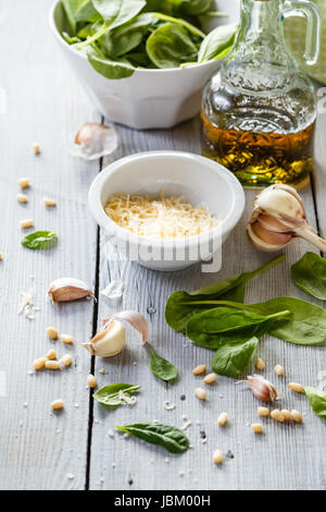 Prodotti per fare in casa il pesto: foglie di spinaci, formaggio grattugiato, aglio, pinoli, olio di oliva e frullatore bianco su sfondo di legno Foto Stock