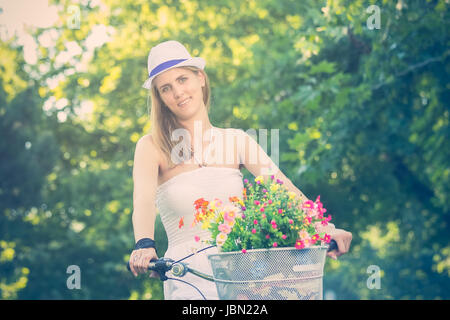 Femmina elegante su vintage hipster bike con cesto pieno di fiori in un parco. Le persone attive, vacanza, lo stile di vita e concetti. In stile retrò immagine. Foto Stock