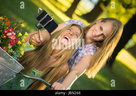 Due belle donne facendo loro foto con lo smartphone utilizzando selfie stick in natura. Divertimento, la gioventù e i concetti di stile di vita. Foto Stock