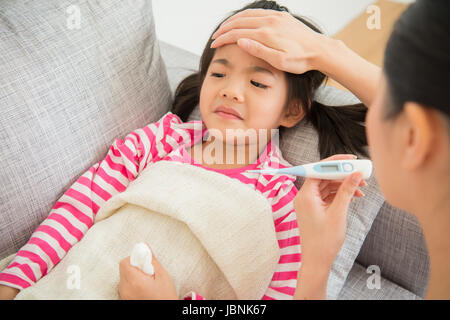 Incantevole piccola ragazza asiatica è malato sdraiato sul divano letto madre la verifica di un termometro e di toccare la figlia di fronte nel salotto di casa. famiglia acti Foto Stock