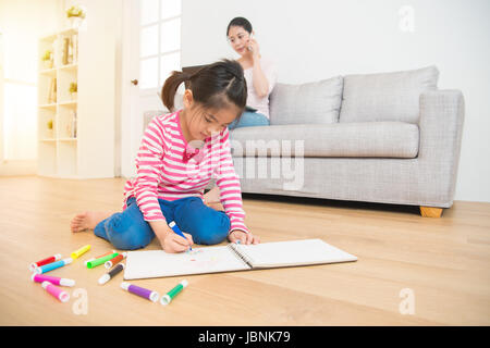 Asian madre cinese a lavorare con il computer a parlare al telefono e bambini ragazza sdraiato sul pavimento in legno disegno sketchbook nel salotto di casa Foto Stock