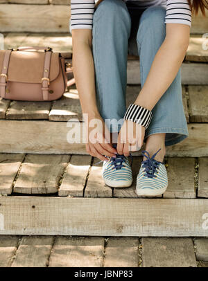 Giovane donna in blue jeans e scarpe da ginnastica a strisce si siede sul vecchio gradini in legno e si lega a lacci delle scarpe. Messa a fuoco selettiva. Foto Stock