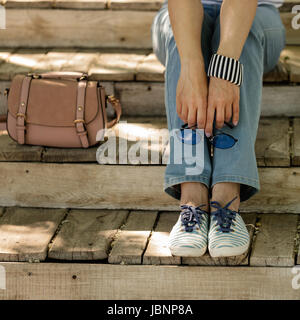 Giovane donna in jeans blu, stripe sneakers siede sul vecchio gradini in legno e tenere gli occhiali da sole. Messa a fuoco selettiva. Foto Stock