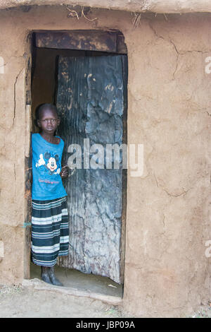 Maasai bambino in piedi nel vano della porta di una capanna di fango, indossando un Mickey Mouse T-shirt, in un villaggio vicino al Masai Mara, Kenya, Africa orientale Foto Stock