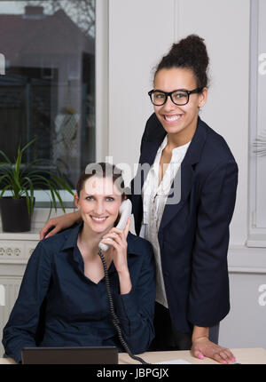 Eine junge, lächelnde Büroangestellte sitzt am Schreibtisch vor einem Notebook und telefoniert. Eine weitere lächelnde Frau steht neben ihr. Foto Stock