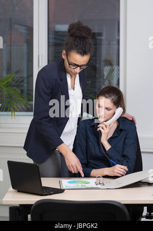 Eine junge Büroangestellte sitzt am Schreibtisch vor einem Aktenordner und telefoniert. Eine weitere Frau steht neben ihr und zeigt auf den Aktenordner. Foto Stock