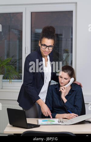 Eine junge Büroangestellte sitzt am Schreibtisch vor einem Aktenordner und telefoniert. Eine weitere Frau steht neben ihr und zeigt auf den Aktenordner. Foto Stock