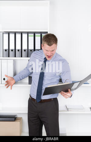 Junger wütender Geschäftsmann steht vor einem Regal im Büro und schaut in einen geöffneten Aktenorder. Foto Stock