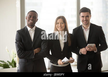 Allegro imprenditrice in abbigliamento formale in piedi fra due imprenditori fiduciosi che posano per una fotocamera, multi-etnico ritratto team motivato e professionale Foto Stock