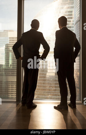 Verticale vista posteriore di afro-americani e imprenditori caucasica nel convenzionale supporto vicino alla grande finestra guardare grande città al di fuori degli edifici, due diverse m Foto Stock