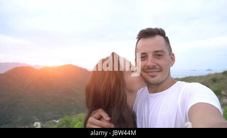 Coppia sorridente tenendo selfie in montagna, antenna vista città. Donna Uomo baci. Foto Stock