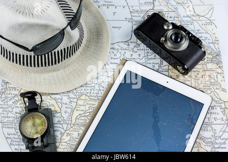 Un'antica mappa, una fotocamera vintage, un tablet con flight tracker, occhiali da sole e un cappello bianco, tutto organizzato per evocare un'avventura di viaggio e un concetto di viaggio Foto Stock