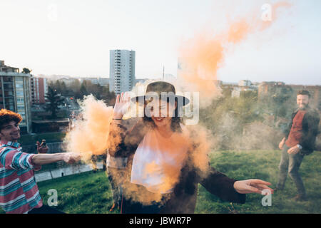 Il gruppo di amici sul tetto, tenendo il fumo colorato flares, giovane donna a piedi attraverso fumo arancione Foto Stock