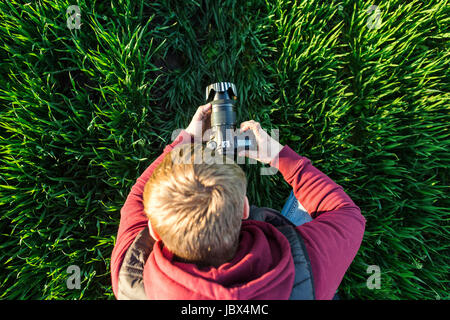 Affascinante la fotografia e il processo di ripresa del concetto - vista superiore del giovane uomo maschio con specchio telecamera in mani in piedi in un campo di luminoso verde erba, jeans e giacca felpa con cappuccio. Foto Stock