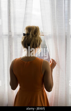 Vista posteriore di una bionda Caucasian woman standing dalla finestra che guarda lontano Foto Stock