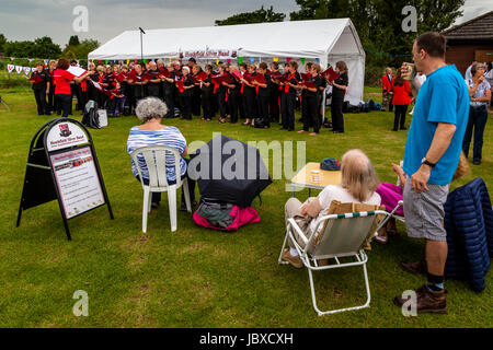 La popolazione locale guarda una comunità coro cantando sotto la pioggia a Maresfield Fete, Maresfield, East Sussex, Regno Unito Foto Stock