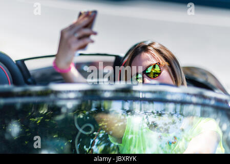Giovane donna alla guida di una cabriolet auto mentre distratti facendo una selfie con il suo smartphone e risultare pericoloso per il traffico intorno a. Foto Stock