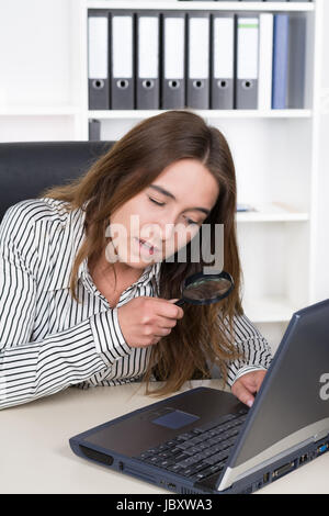 Eine junge Frau schaut durch eine Lupe auf ein Laptop am Schreibtisch im Büro. Im Hintergrund ist ein regale. Die Frau schaut zum Notebook. Foto Stock