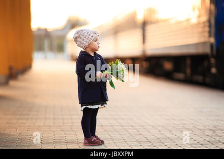 Ragazza con i fiori presso la stazione ferroviaria. Kid attendere per mom sulla luce del tramonto sul treno in arrivo sullo sfondo. Foto Stock