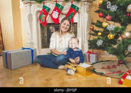 Felice giovane madre seduta con il suo bambino figlio sul pavimento nel salone accanto al caminetto e albero di natale Foto Stock