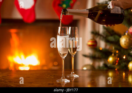 Persona che riempie due bicchieri di champagne. La masterizzazione di camino e albero di Natale decorato sullo sfondo Foto Stock