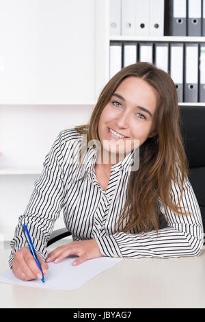 Eine junge, lächelnde Frau schreibt Mit einem blauem Stift auf einem Blatt Papier am Schreibtisch im Büro. Im Hintergrund steht ein regale. Die Frau schaut zur Kamera. Foto Stock