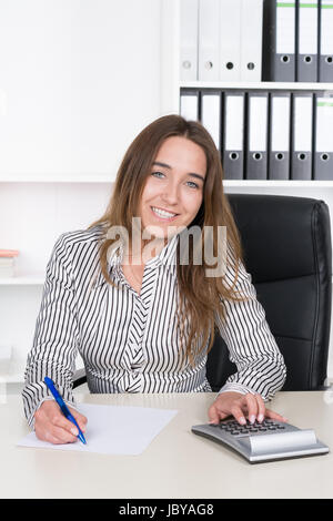 Eine junge, lächelnde Frau"führt Mit einem Tischrechner Berechnungen durch und macht Notizen sich mit einem blauem Stift auf einem Blatt Papier am Schreibtisch im Büro. Im Hintergrund steht ein regale. Die Frau schaut zur Kamera. Foto Stock