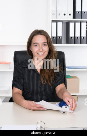 Eine junge, lächelnde Frau locht Dokumente am Schreibtisch im Büro. Im Hintergrund steht ein regale. Die Frau schaut zur Kamera. Foto Stock