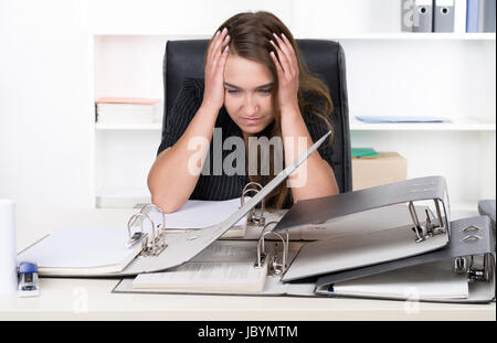 Eine junge, frustrierte Frau sitzt vor einem Haufen von Akten am Schreibtisch im Büro. Im Hintergrund steht ein regale. Die Frau schaut nach unten. Foto Stock