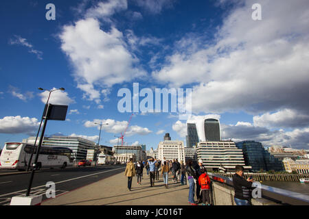 I turisti e la gente del posto si mescolano sul London Bridge sul fiume Tamigi nel sole sotto un cielo drammatico con scudding nuvole riflettono nel ponte ferrovia Foto Stock