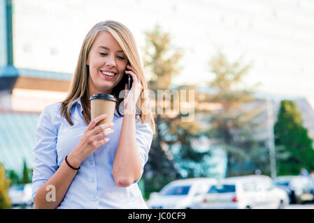 Close up ritratto di una gioiosa imprenditrice usando il suo smartphone e portante le cartelle di lavoro in piedi contro un soleggiato blue sky. Foto Stock