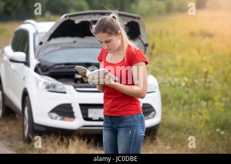 Ritratto di giovane donna ha sottolineato in piedi alla vettura rotto e lettura manuale del proprietario Foto Stock