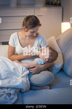 Ritratto di giovane madre premurosa dando una bottiglia con latte per il suo bambino a letto a notte. Concetto di Parenting Foto Stock