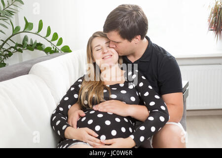 Ritratto di uomo bello seduto sul divano e baciare moglie incinta nella guancia Foto Stock
