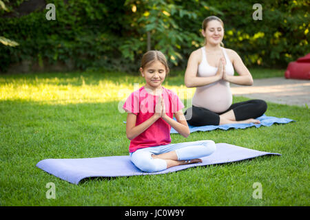 Giovane donna incinta e carina ragazza a praticare yoga su erba a park Foto Stock