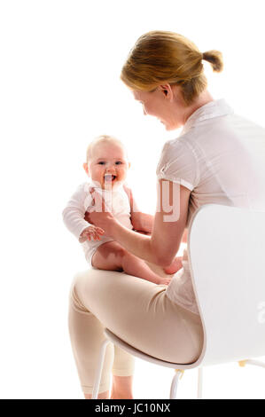 Erwachsene Frau sitzt auf einem weissen Stuhl und das haelt 6 Monate alte Baby mit beiden Haenden fest, vor isoliert weissem Hintergrund. Foto Stock