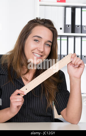 Eine junge, lächelnde Frau hält ein Lineal aus Holz am Schreibtisch im Büro. Im Hintergrund steht ein regale. Foto Stock
