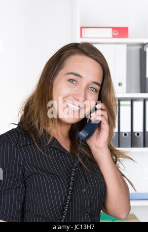 Eine junge, lächelnde Frau telefoniert im Büro. Im Hintergrund steht ein regale. Die Frau schaut zur Kamera. Foto Stock
