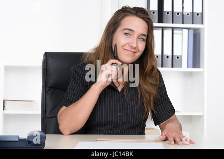 Eine junge, lächelnde Frau hält einen Stift un ihre Wange während sie am Schreibtisch im Büro sitzt. Im Hintergrund steht ein regale. Foto Stock