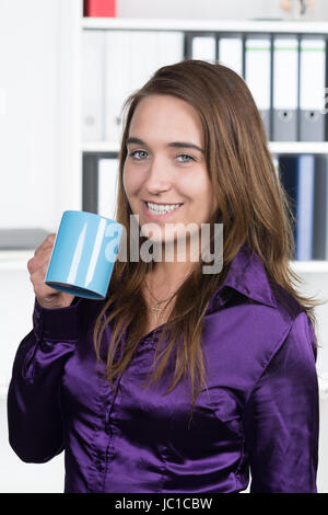 Eine junge, lächelnde Frau hält eine blaue Tasse Kaffee während sie im Büro steht. Im Hintergrund befindet sich ein regale. Die Frau schaut zur Kamera. Foto Stock