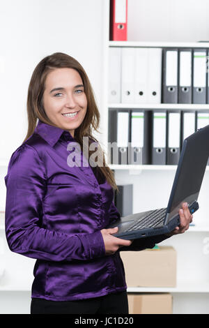 Eine junge, lächelnde Frau hält ein Laptop während sie im Büro steht. Im Hintergrund befindet sich ein regale. Die Frau schaut zur Kamera. Foto Stock